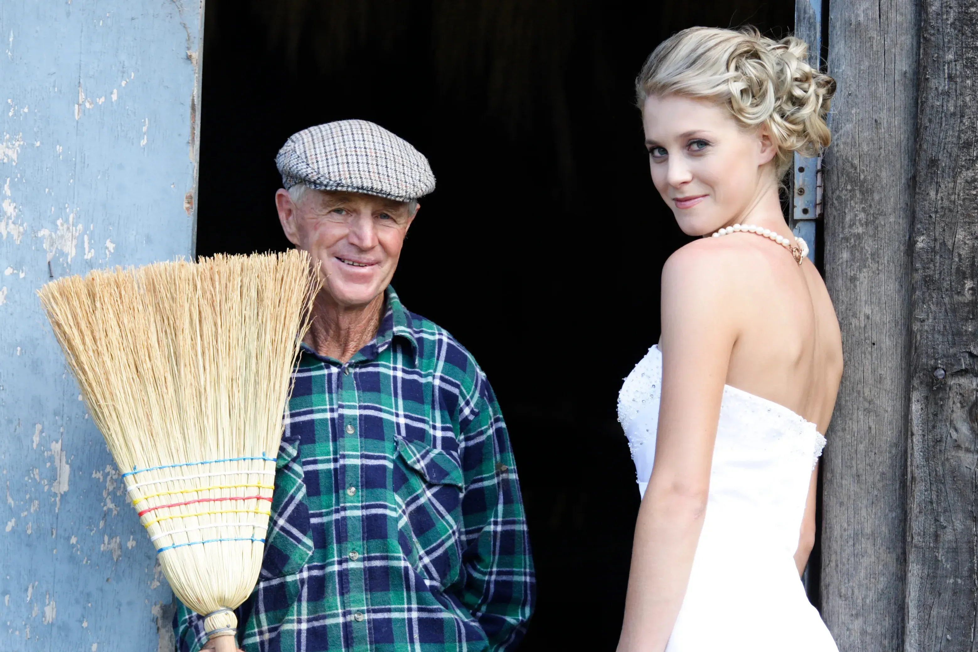 A bride in a barn doorway with a farmer holding a broom, her formal updo styled by Amanda Frost