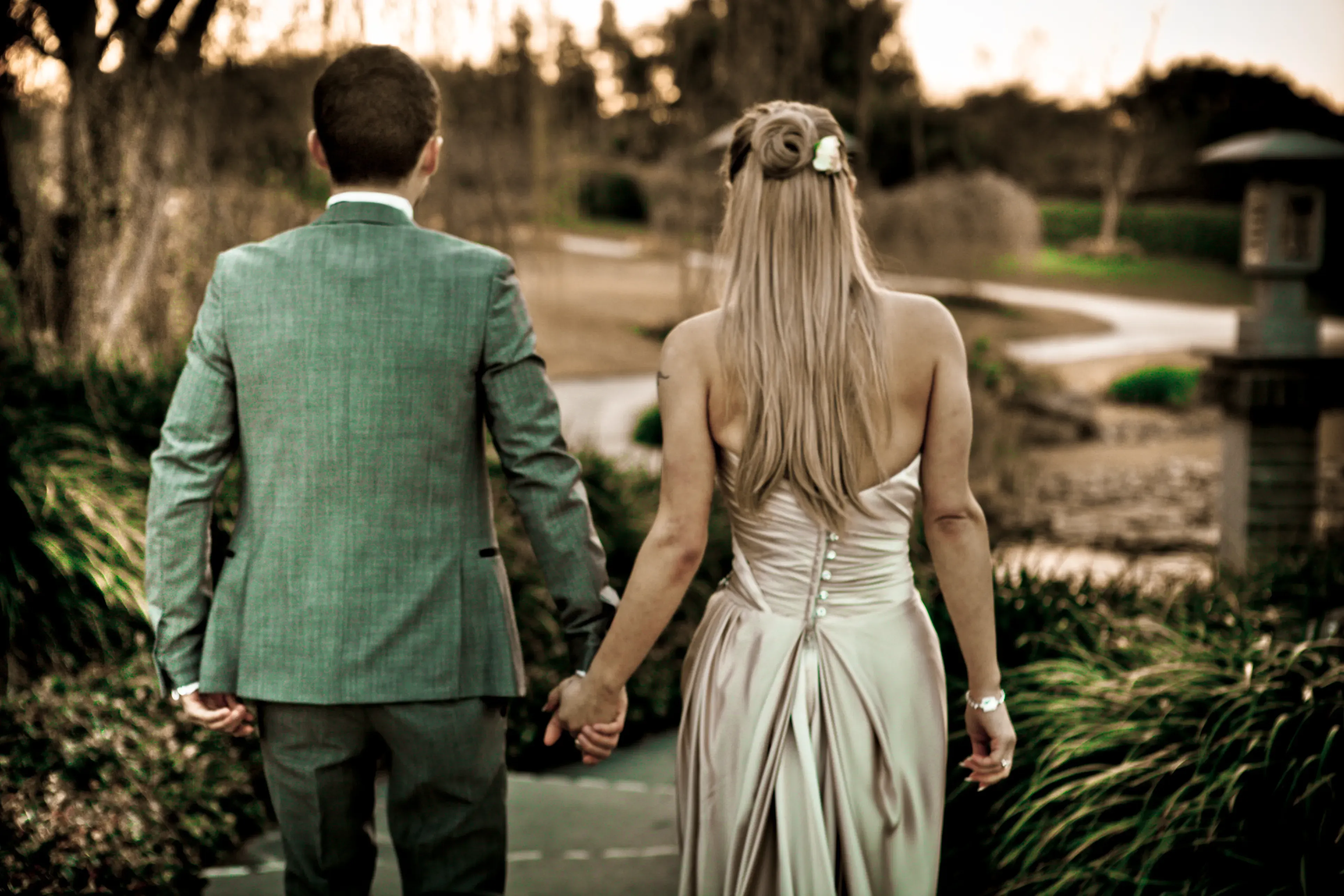A bride and groom walking hand in hand through a garden at sunset, the bride’s hair styled in a half-up twist by Amanda Frost